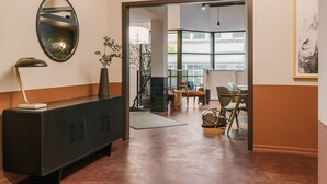 Un hall d’hôtel moderne avec un meuble d’entrée en bois foncé, un grand miroir et une vue sur un espace de restauration avec des tables et des chaises.