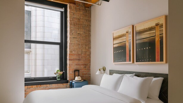 Une chambre d’hôtel avec un grand lit, une fenêtre donnant sur des immeubles, une table de chevet avec une lampe et deux tableaux encadrés au mur.