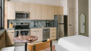 Une cuisine compacte équipée d’un four à micro-ondes, d’une cuisinière et d’un réfrigérateur, une petite table à manger avec une chaise, et un lit dans la pièce adjacente.