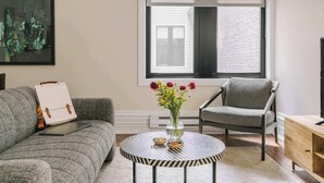 Un salon moderne avec un canapé gris, une table basse ronde recouverte d’une nappe à rayures, un fauteuil gris et un meuble en bois.