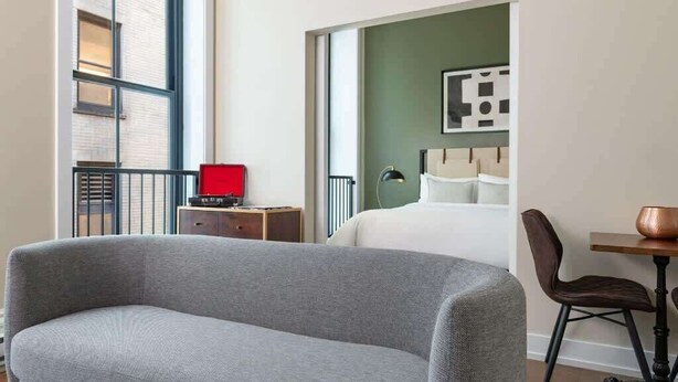 Een hotelkamer met een grote grijze bank, een tafeltje met een koperen pot, een rode lamp en een bed met witte lakens.