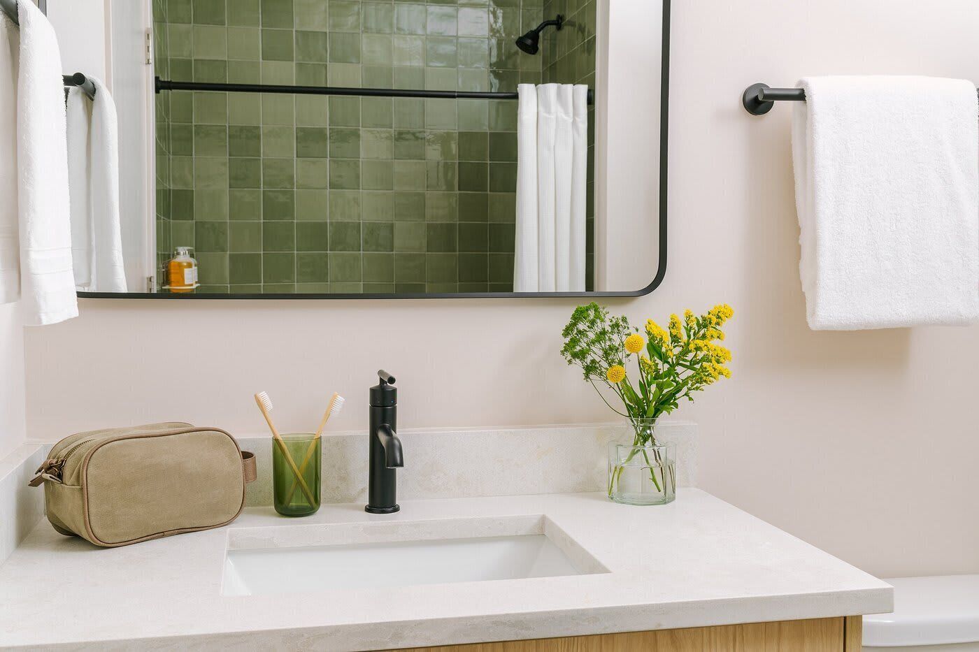 Un bagno con un lavandino bianco, uno specchio, un portasciugamani, un vaso di vetro con fiori gialli e una borsa beige sul ripiano.