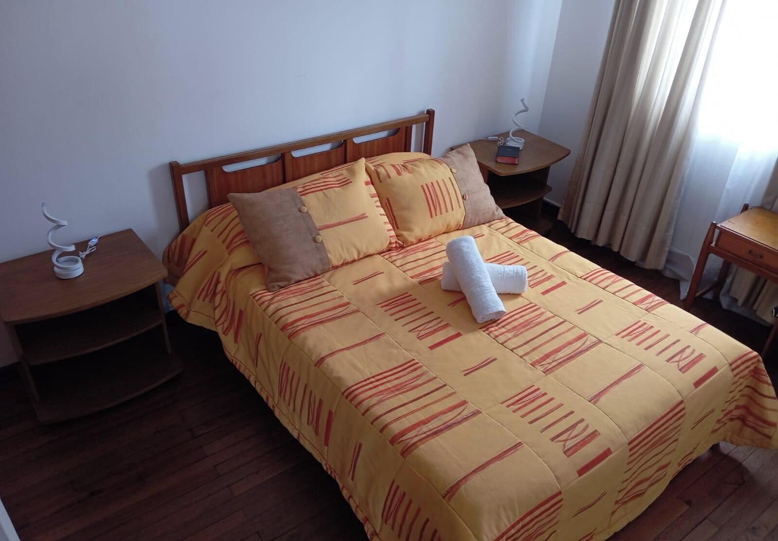 Une chambre d’hôtel avec un lit en bois, une literie jaune, deux oreillers, une table de chevet avec une lampe et une fenêtre avec des rideaux.