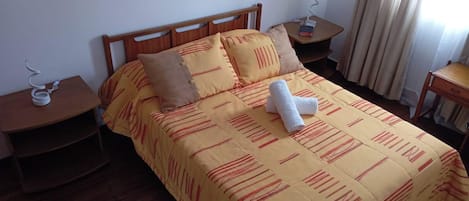 Ein Hotelzimmer mit einem hölzernen Bett, gelber Bettwäsche, zwei Kissen, einem Nachttisch mit Lampe und einem Fenster mit Vorhängen.