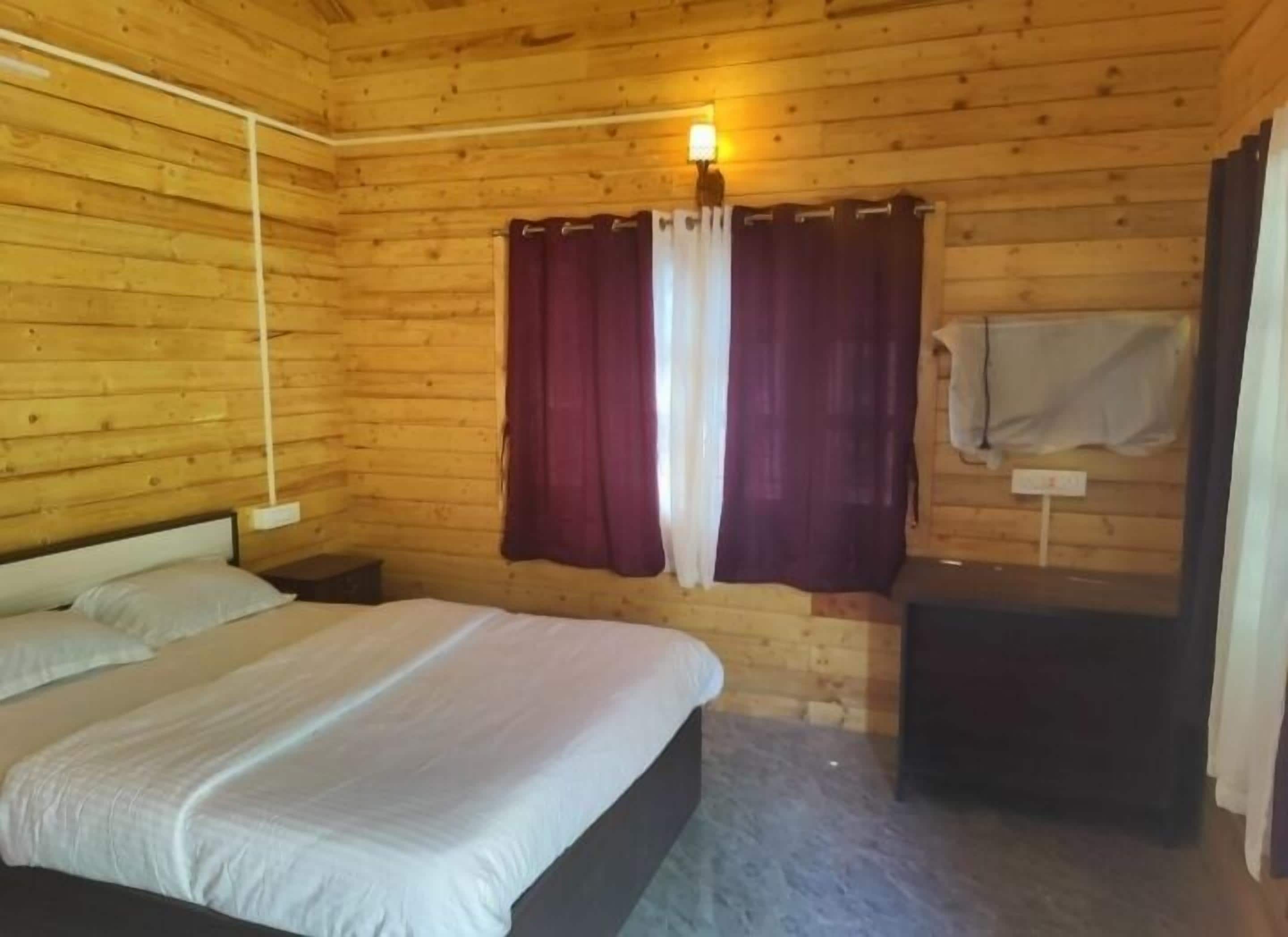 Une chambre dans une cabane en bois, avec un lit, une table de chevet, une lampe fixée au mur et une fenêtre avec des rideaux.