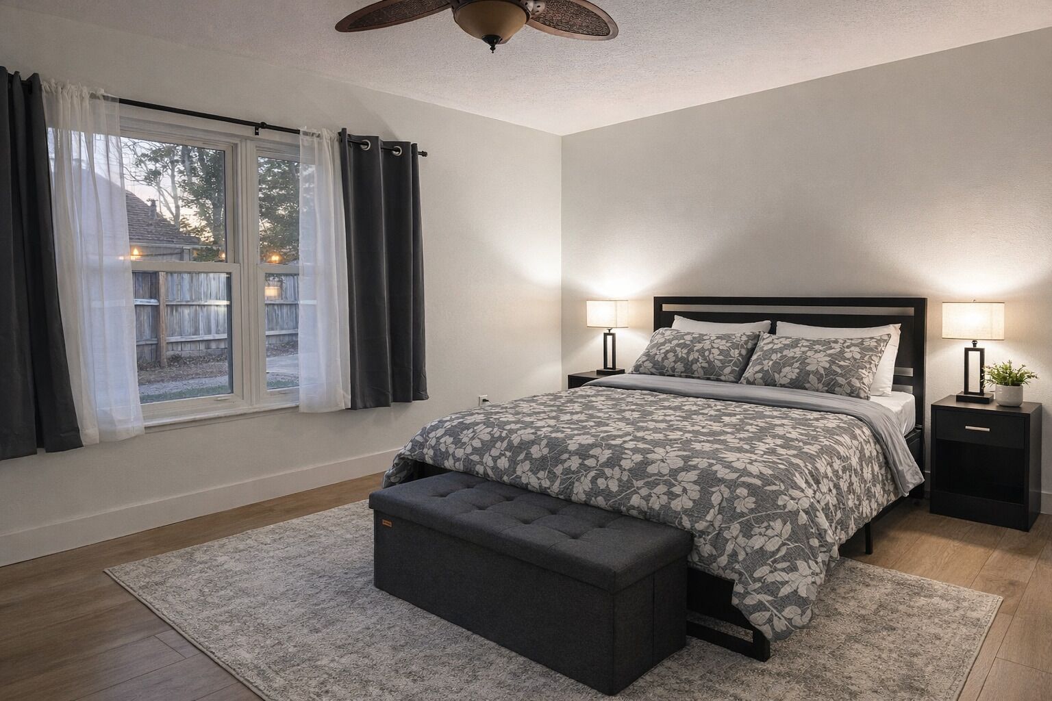 A bedroom with a bed, bedside tables, a bench, a ceiling fan, and a window with curtains.