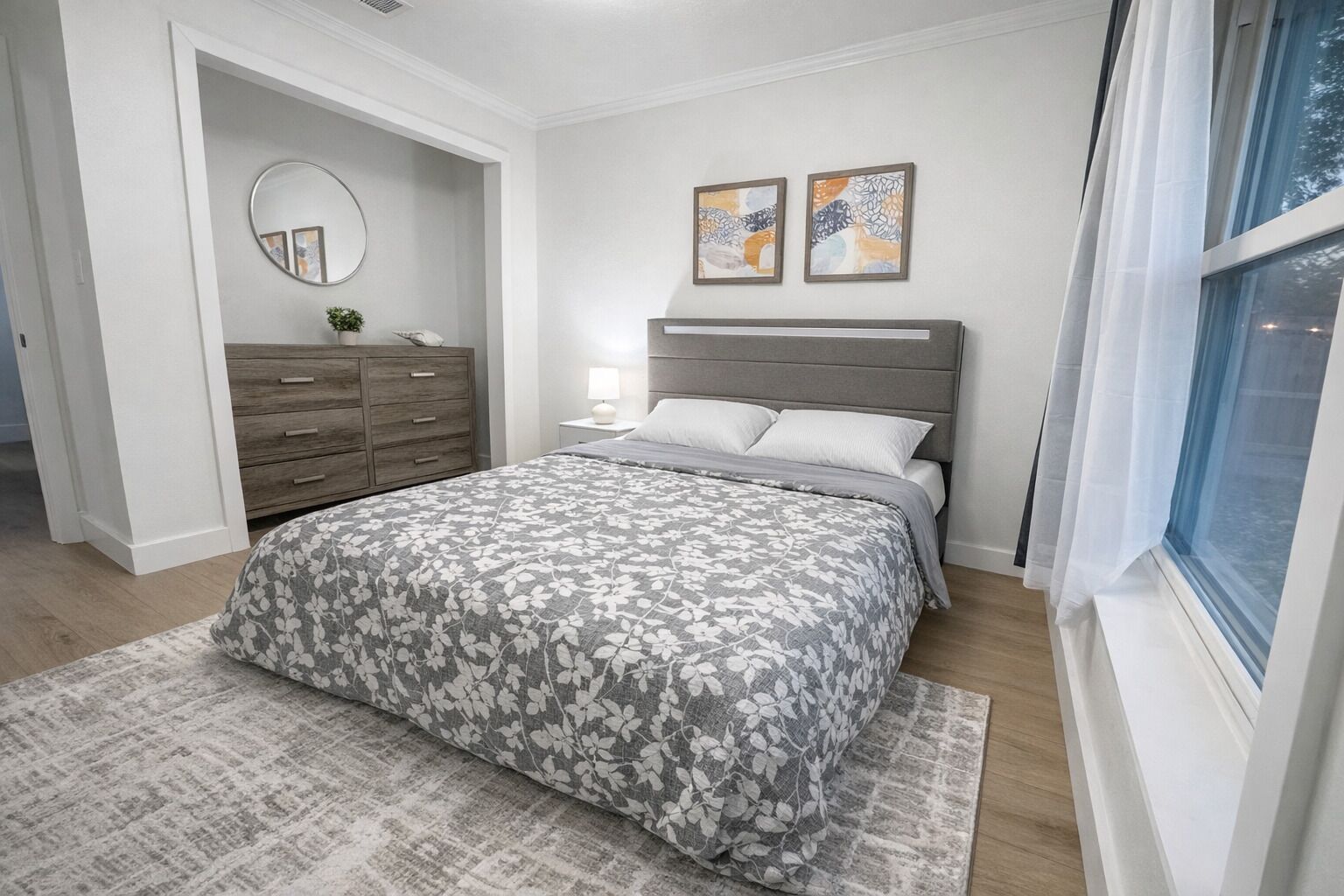 Un dormitorio con cama, mesita de noche, cómoda, espejo y un cuadro abstracto en la pared.