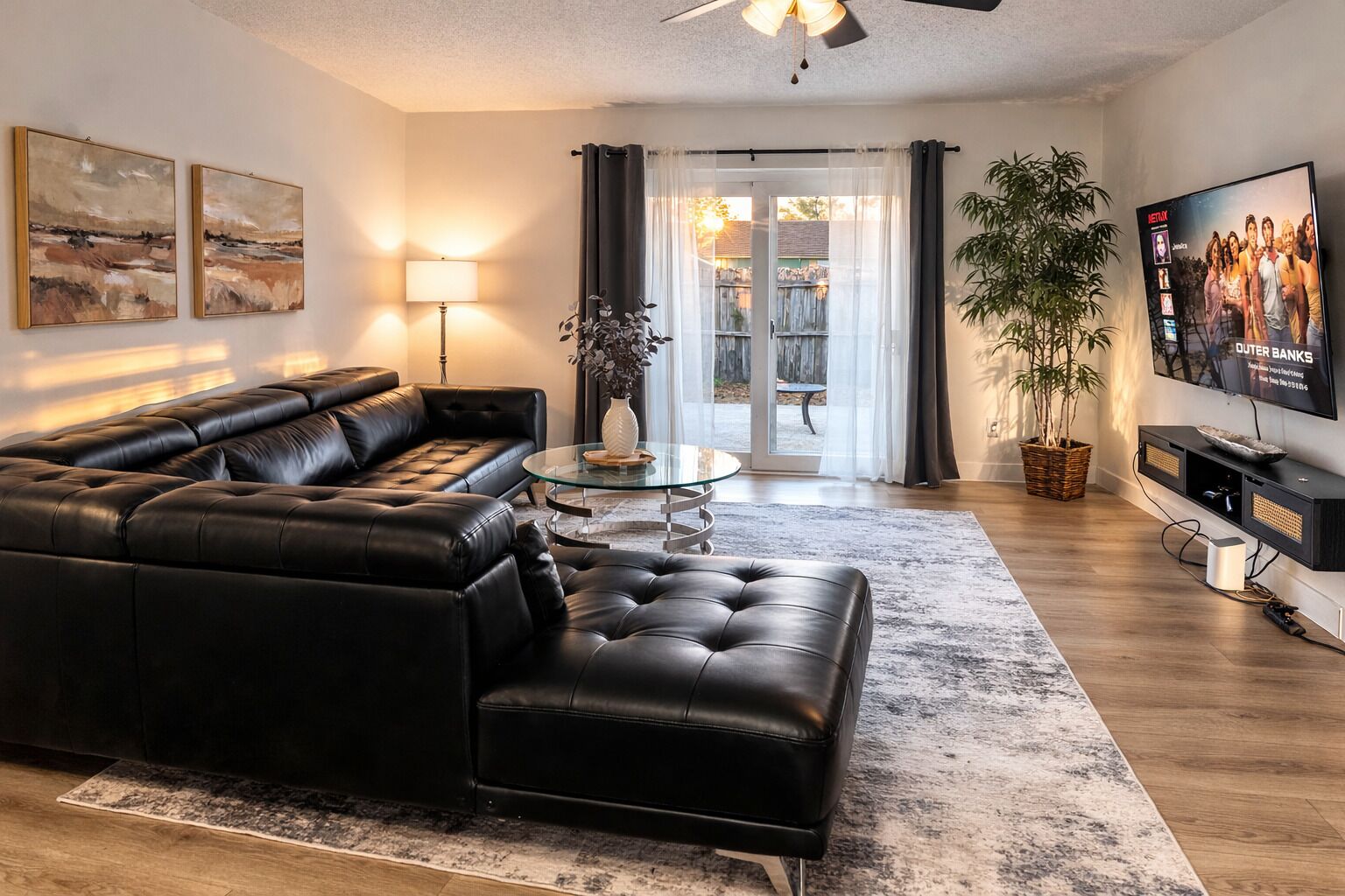 A modern living room with a black leather sectional sofa, a glass coffee table, and a flat-screen TV mounted on the wall.