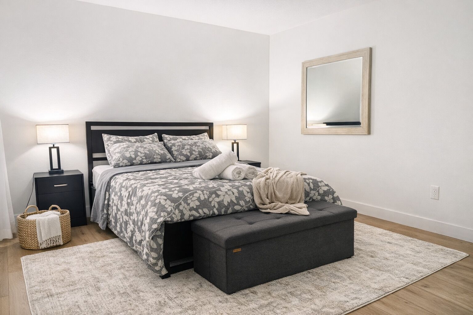 Un dormitorio con cama, mesitas de noche, un espejo, una lámpara de noche, una cesta tejida y un otoman gris.