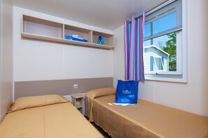 Een hotelkamer met twee enkele bedden, een klein tafeltje, een plank met decoratieve spullen en een raam met gordijnen.
