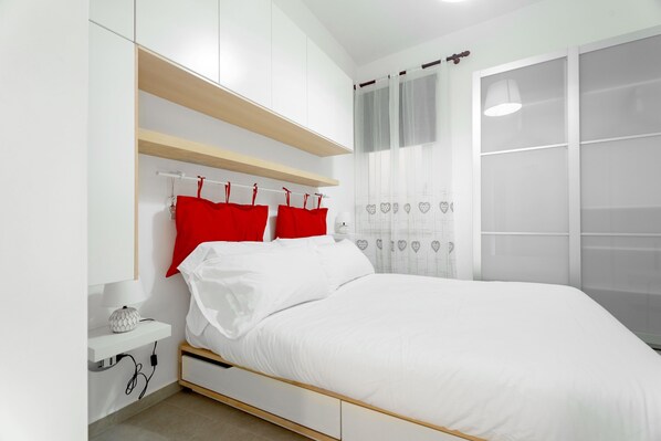 A bedroom with a bed, white bedding, red pillows, a bedside table with a lamp, and a window with sheer curtains. - Heart in Home - One Bedroom Apartment, Sleeps 2 (Monopoli)