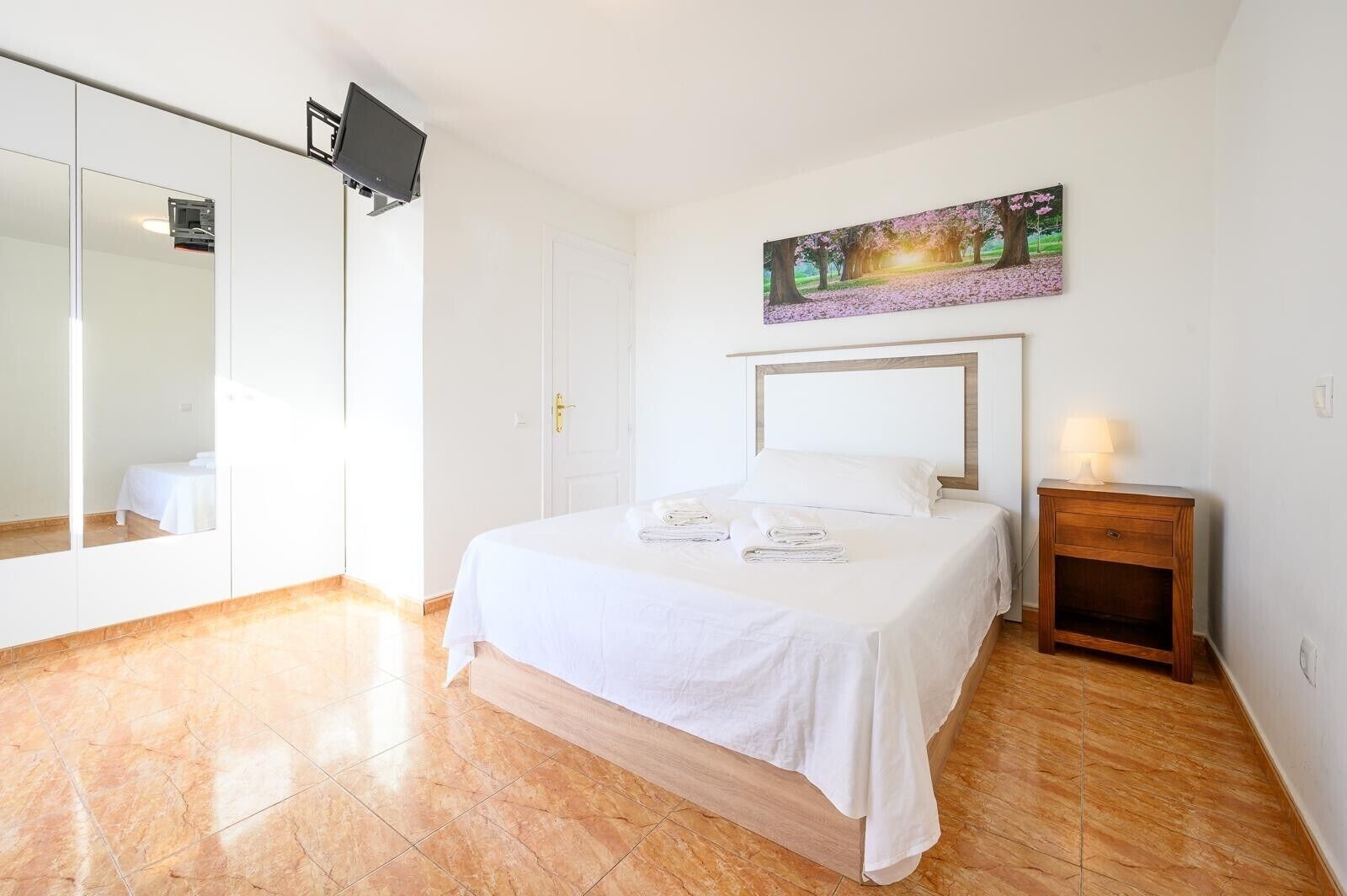 A bedroom with a bed, a bedside table with a lamp, a painting on the wall, and a large mirror.