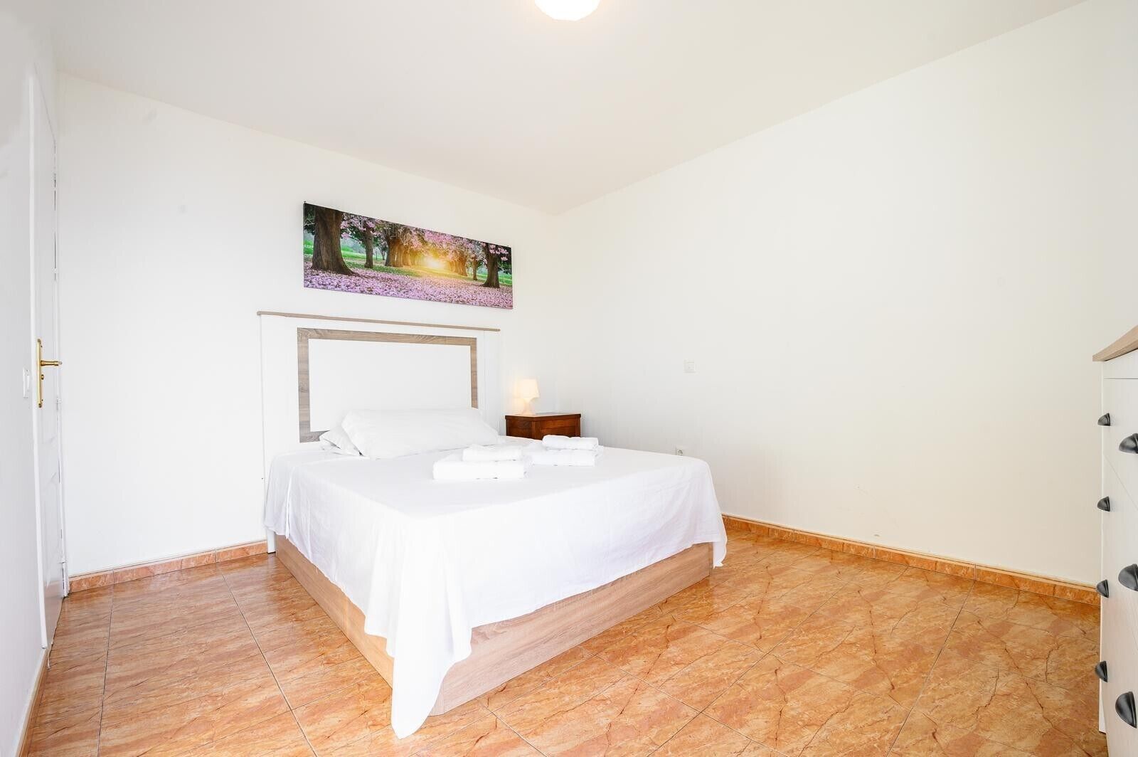 Quarto com cama, mesa de cabeceira, quadro acima da cama e porta à esquerda.