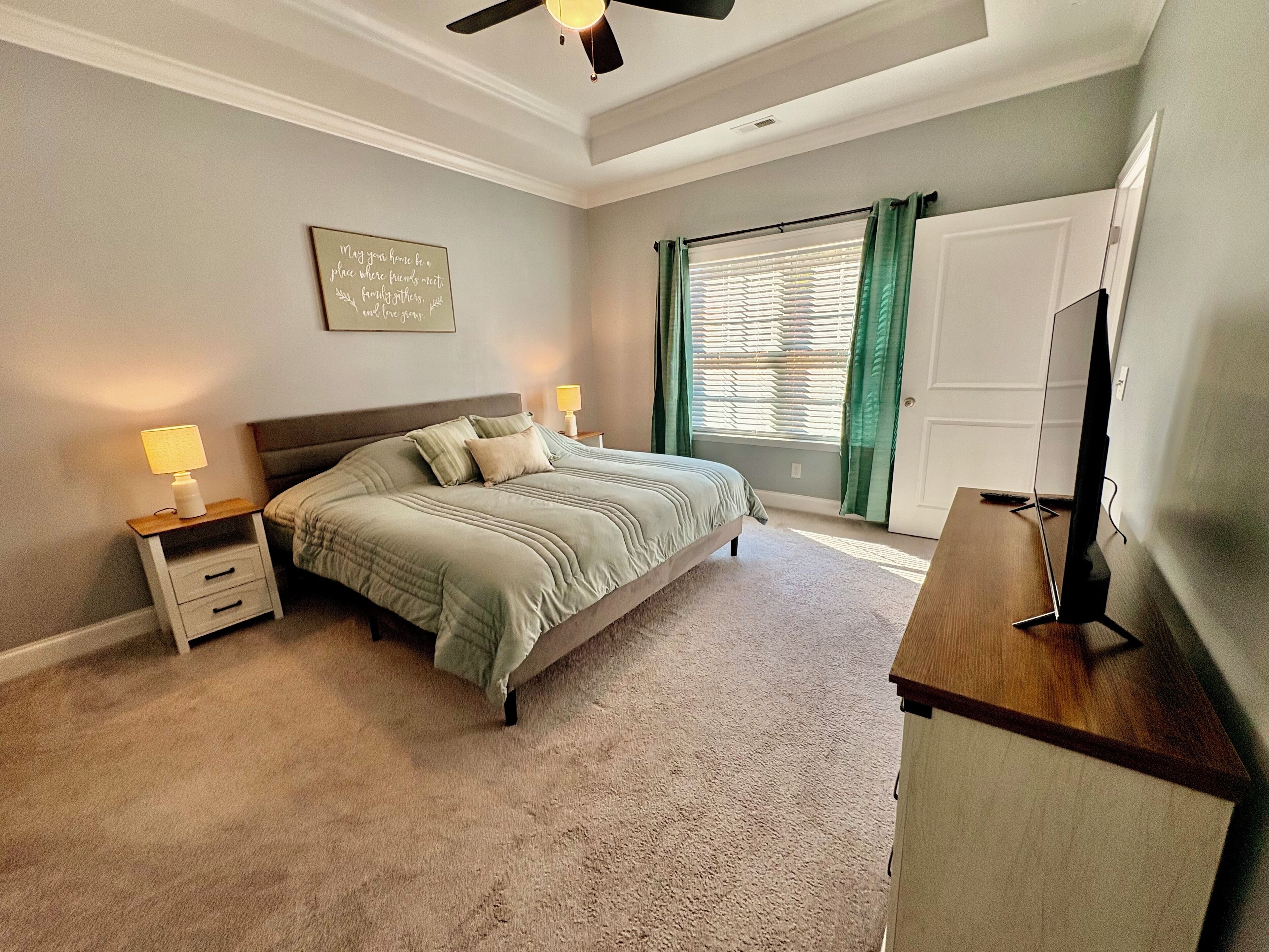 A bedroom with a bed, nightstands, a desk, a ceiling fan, a window with curtains, and a door.