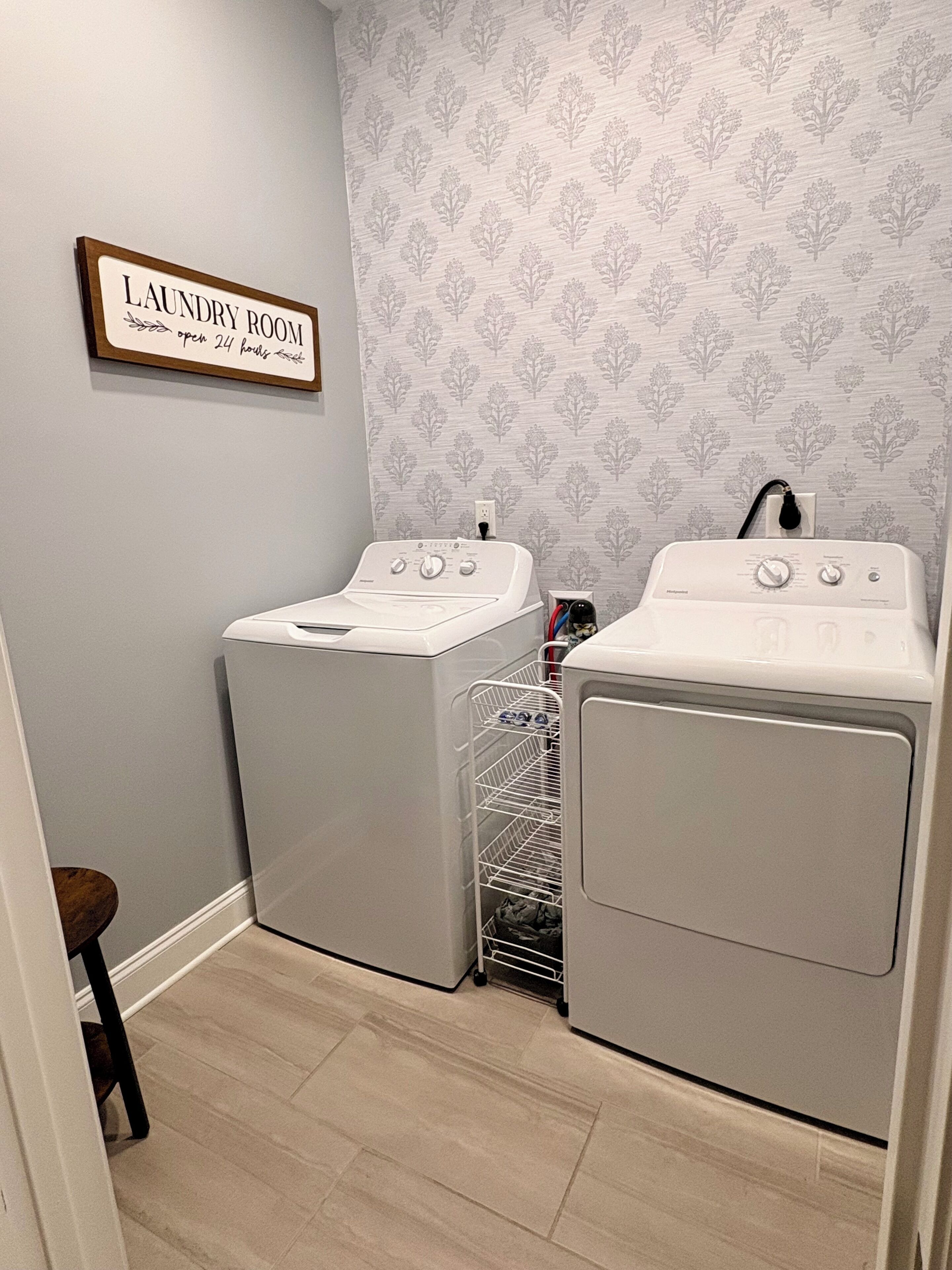 Une buanderie équipée de deux machines à laver et de deux sèche-linge à chargement frontal, tous deux de couleur blanche, d’un étendoir métallique et d’un panneau indiquant qu’elle est ouverte 24 heures sur 24.
