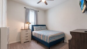 Une chambre à coucher avec un lit, une commode, un ventilateur de plafond et un téléviseur fixé au mur.