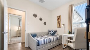 Un espace de vie compact comprenant un canapé, un bureau et une chaise, une fenêtre avec des stores et une porte donnant sur une autre pièce.