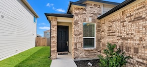 A residential building with a brick facade, a black door, and a small front yard with a welcome mat. - Modern Retreat near RandolphAFB- Family Friendly, Coffee Bar, WiFi, Yard &Garage (Converse)
