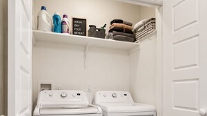 Une buanderie équipée de deux machines à laver et de deux sèche-linge à chargement frontal, tous deux de couleur blanche, d’une étagère contenant des produits de lessive et d’un plancher en bois.