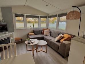 A modern living room with a sectional sofa, two round tables, a television, and large windows offering a view of the outdoors. - Family friendly chalet near the beach on 5* vacation park De Kuilart in Koudum (Koudum)