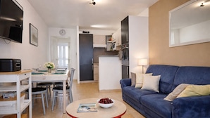 En moderne stue med blå sofa, spisebord med stole, et køkken med skabe og hårde hvidevarer, samt et TV ophængt på væggen.