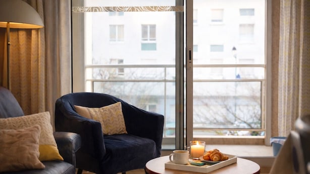 En hyggelig stue med en mørkeblå lænestol, en lædersofa og et rundt træbord med et lys, en kop og et fad med croissanter.