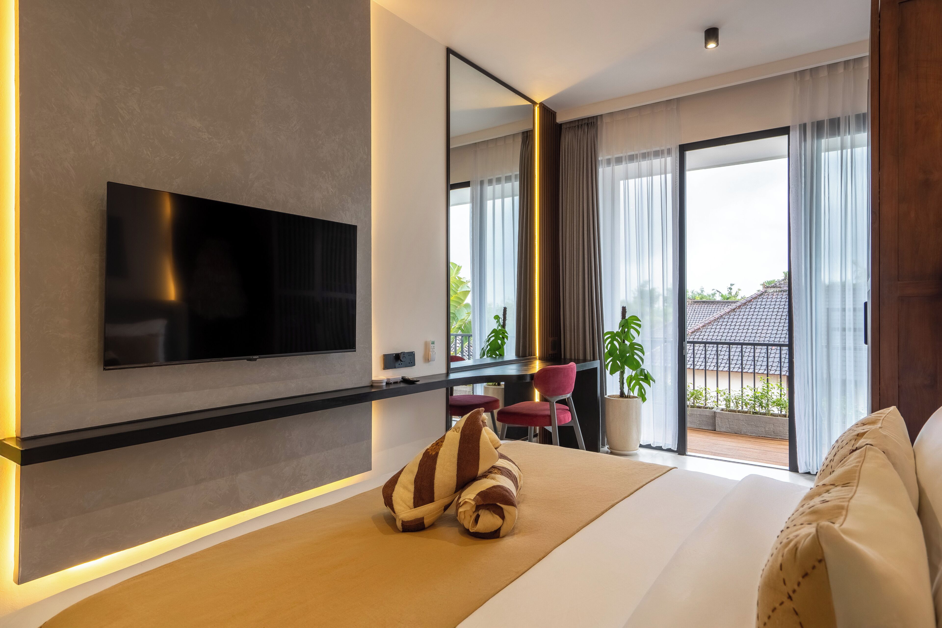A modern hotel room with a large bed, a flat-screen TV mounted on the wall, a desk with a chair, and a balcony with a view of the city.