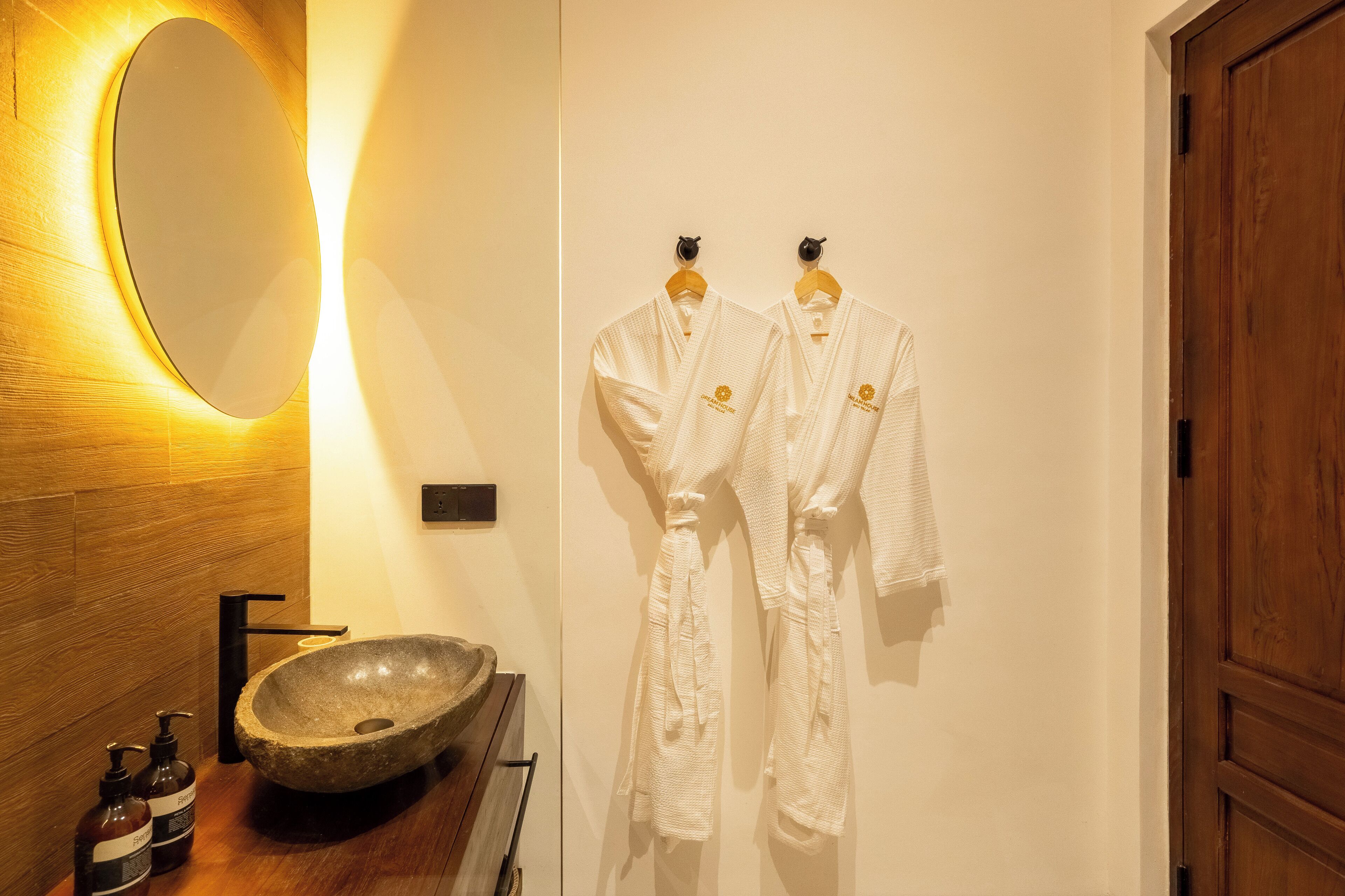 Ein Badezimmer mit einem Holzkosmetikschrank, einem Steintonne, zwei Shampoo-Flaschen und zwei weißen Bademänteln, die an Haken hängen.
