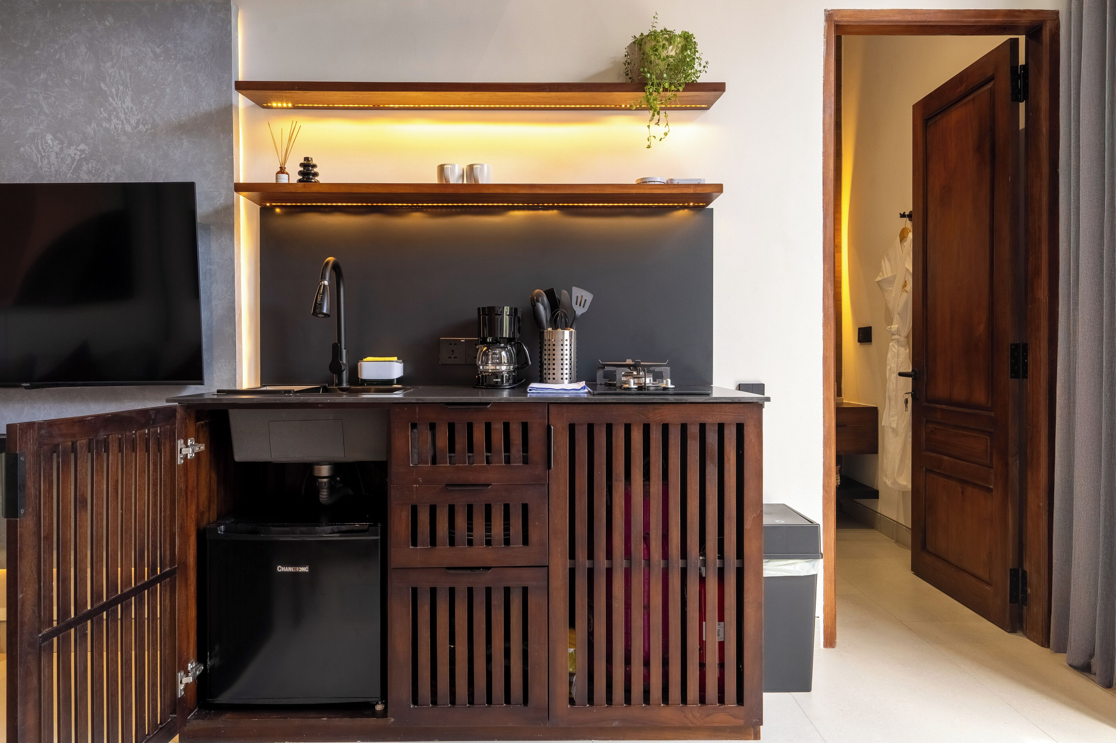 A compact kitchen with a built-in coffee maker, a small refrigerator, and a wooden cabinet with a sliding door.
