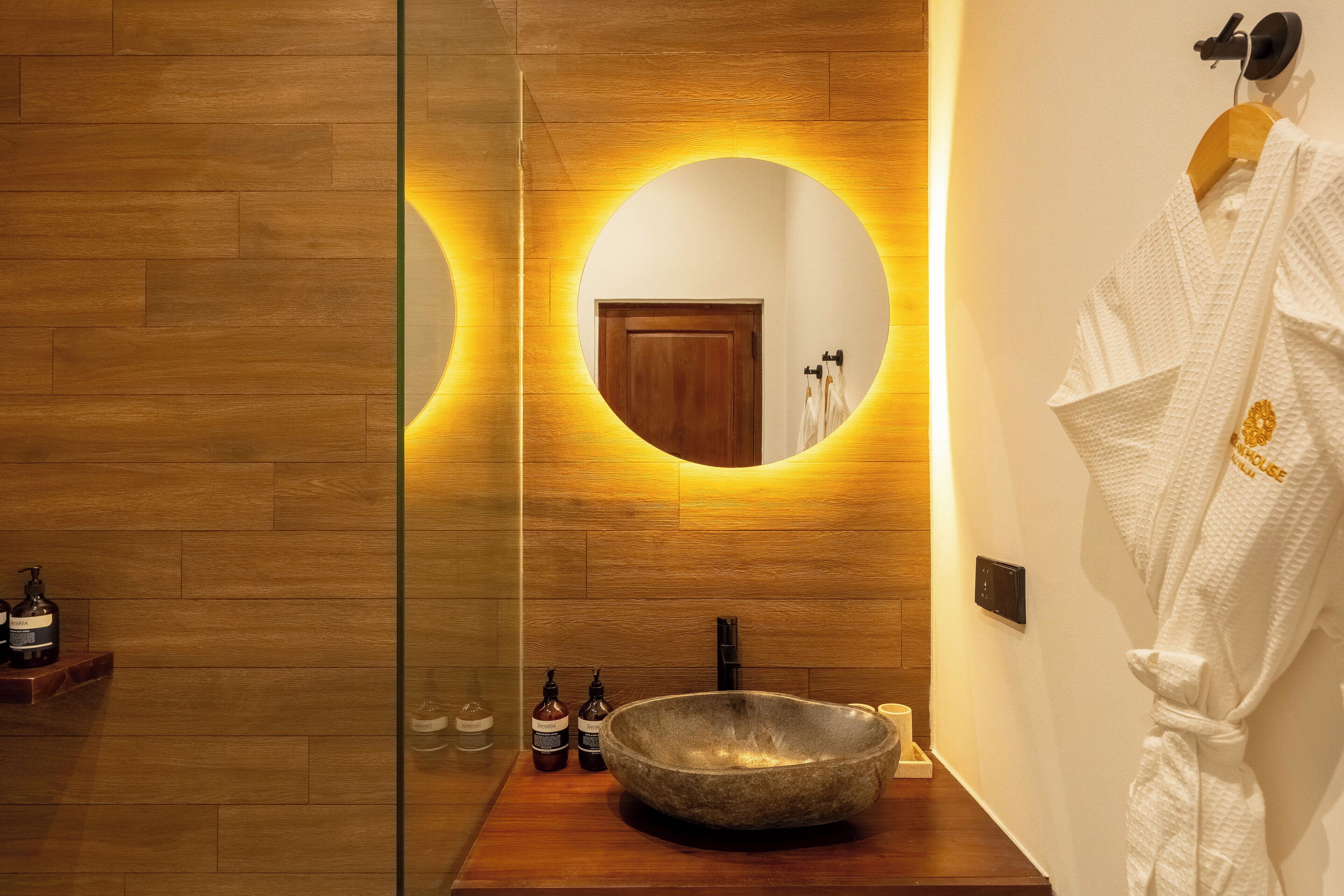 Une salle de bain moderne avec un plancher en bois, un grand miroir rond diffusant une lumière chaleureuse, un vaste lavabo en pierre et une robe blanche accrochée au mur.