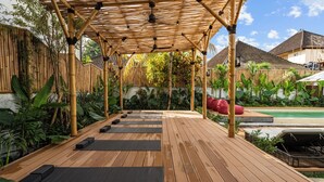 A wooden deck with exercise mats, a bamboo pergola, and a pool in the background. - Palermo Suites by DH (Ubud)