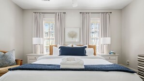 A neatly made bed with a blue blanket and a folded towel, two bedside lamps, a framed picture on the wall, and a window with curtains. - Picture Perfect Charleston Living on Spring St (Charleston)