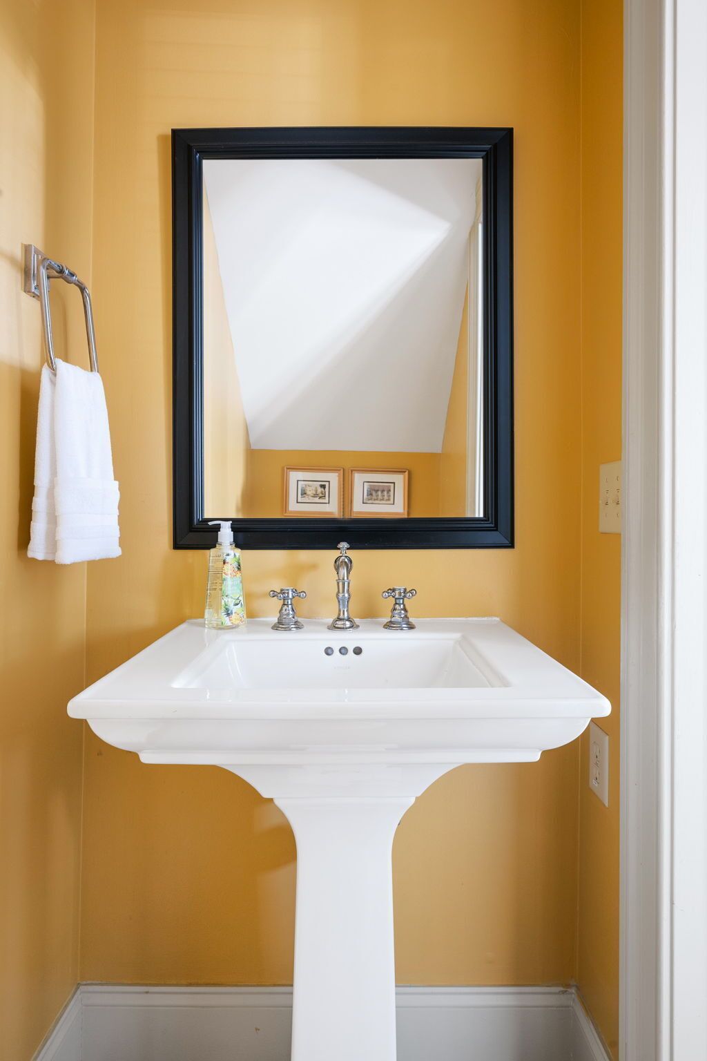 A bathroom with a white pedestal sink, a black-framed mirror, and two framed pictures on the wall.