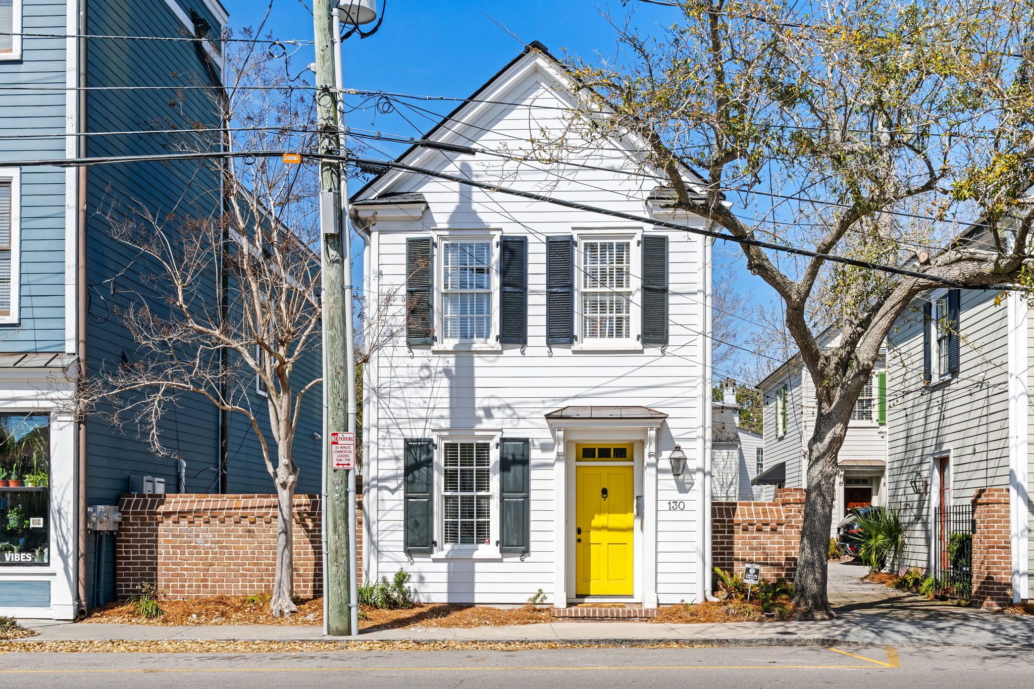 A two-story white house with a yellow door, a brick wall, and a tree in front.