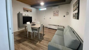 Moderni olohuone, jossa on harmaa sohva, ruokapöytä tuoleineen sekä keittiöalue, jossa on televisio ja kaapistot.