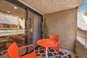 A small balcony with a glass door, a potted plant, and orange chairs and table. - Atomic Starburst – Mountain View Downtown Condo (Palm Springs)