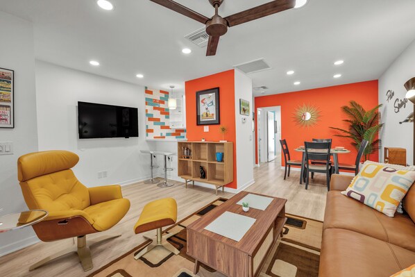 A modern living room with a yellow armchair, a wooden coffee table, and a brown sofa. There is a dining area with a table and chairs, and a kitchen area with a TV and a bookshelf. - Atomic Starburst – Mountain View Downtown Condo (Palm Springs)