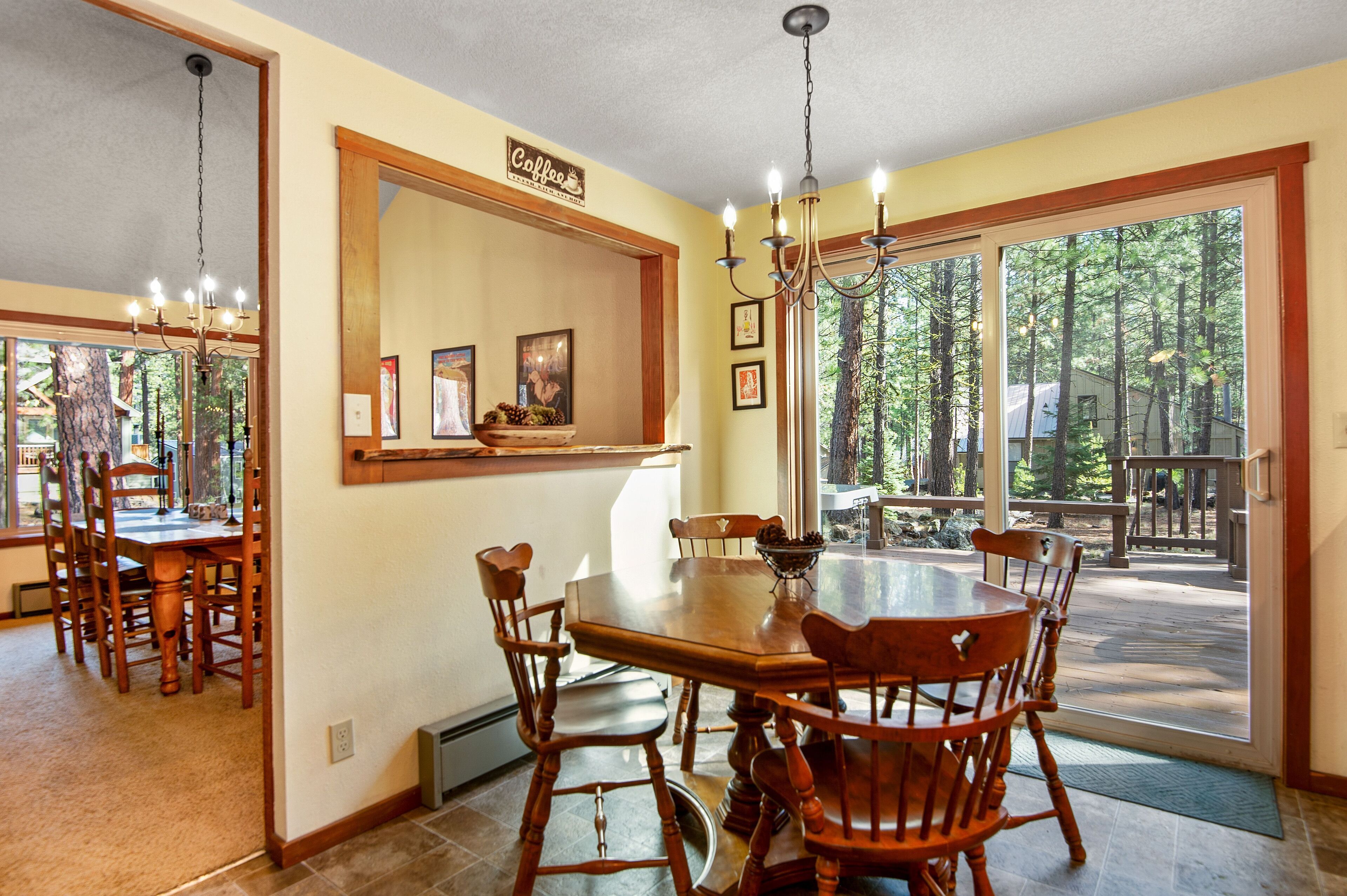 A dining area with a large wooden table and chairs, a chandelier, and a view of a deck and trees through large windows.