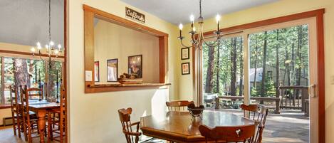 A dining area with a large wooden table and chairs, a chandelier, and a view of a deck and trees through large windows.