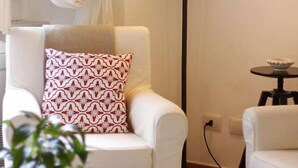 Ein gemütliches Wohnzimmer mit einem weißen Sessel, einem gemusterten Kissen, einer Stehlampe und einem kleinen Tisch mit Zitronen.
