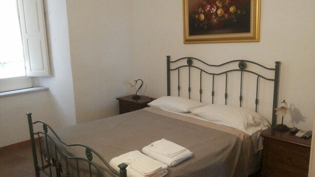 Ein ordentlich bezogenes Bett mit weißen Leinen und einem gefalteten Handtuch, ein Nachttisch mit Lampe und ein Blumenbild an der Wand.