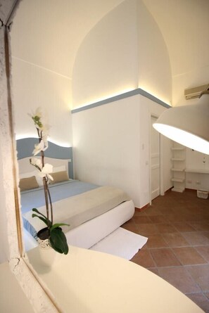 A bedroom with a bed, a small potted plant, a white wall, and a ceiling light.
