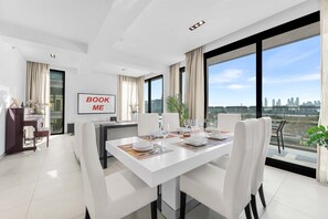 A modern dining area with a white table set for six, large windows offering a city view, and a 'BOOK ME' sign on the wall. - Stayshort - Luxury 3BR Apartment in Meydan That Sleeps 5 (Dubai)