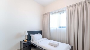 Una cama bien hecha con edredón blanco y una toalla enrollada, una mesita de noche con lámpara, y una ventana con cortinas.