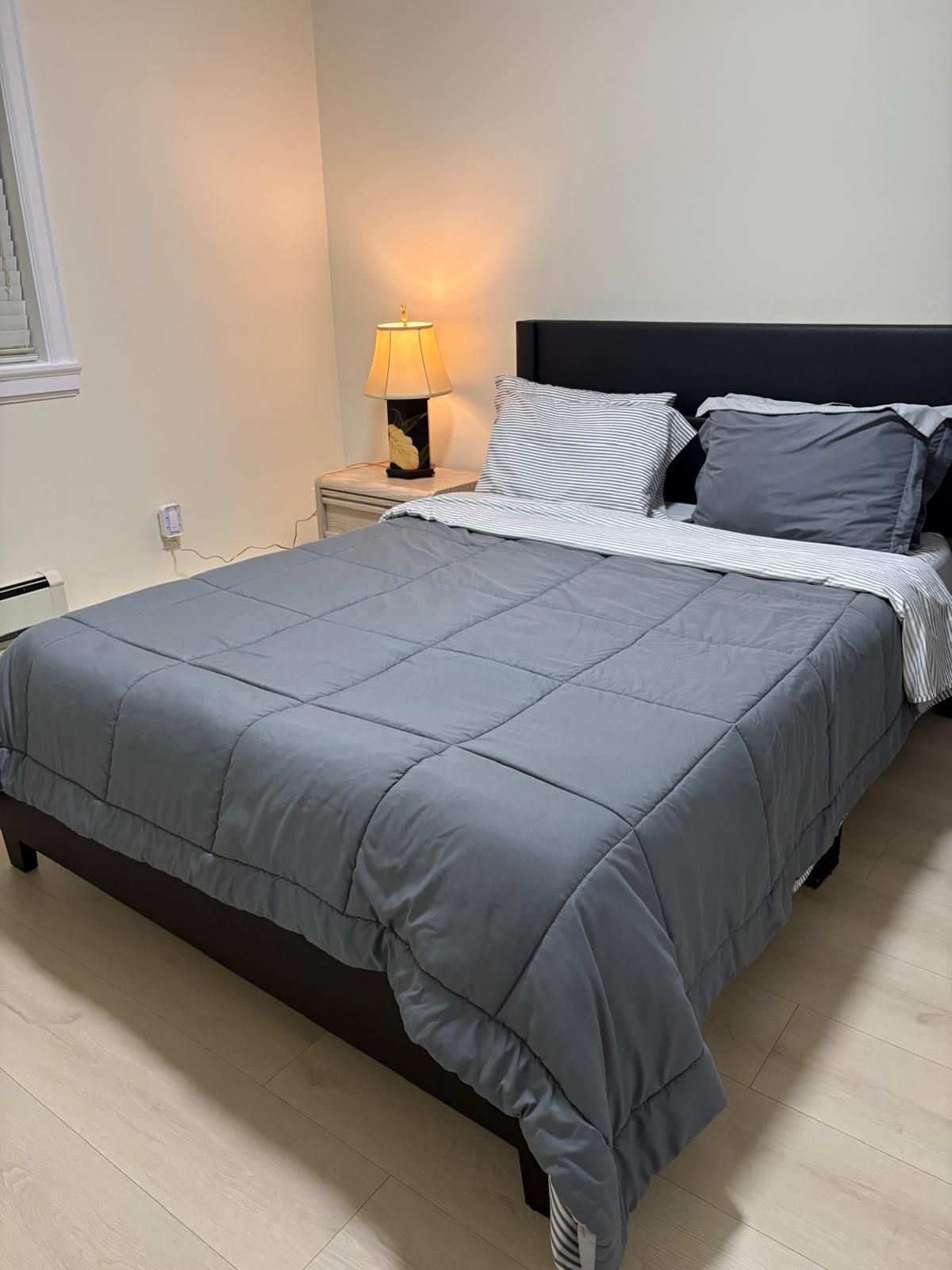 Een bed met een grijze deken, een bedlampje en een nachtkastje met een stopcontact.