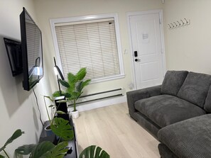 Een woonkamer met een grijze bank, een wandtelevisie, een kamerplant en een raam met zonwering.