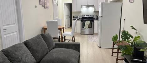 En kompakt opholdsstue med sofa, spisebord, stole, et tekøkken og et tv-bord med en plante.