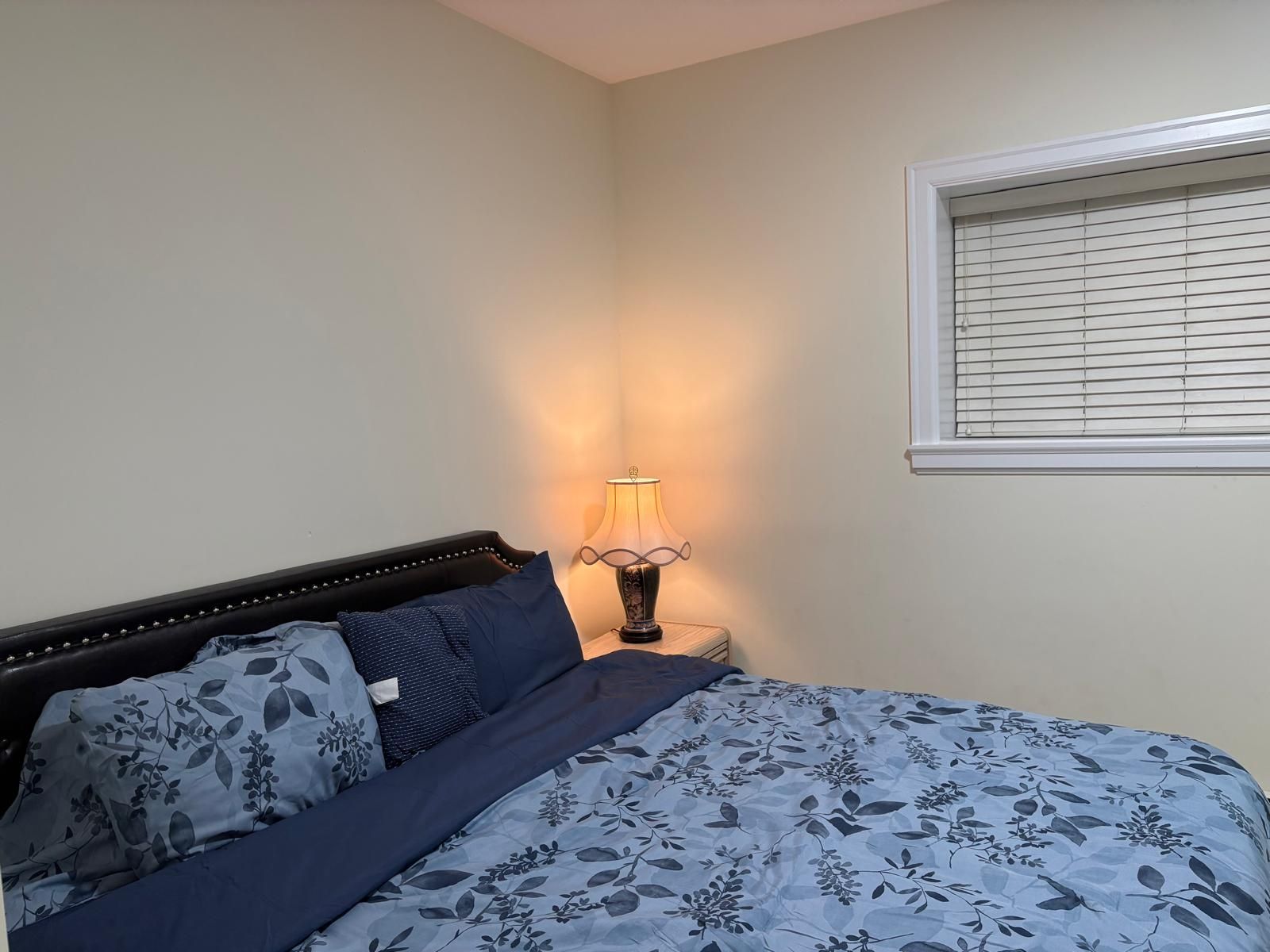 A bedroom with a bed, a bedside lamp, and a window with blinds.