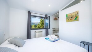 Un dormitorio con una cama, una ventana, un radiador, un cuadro y una mesita.