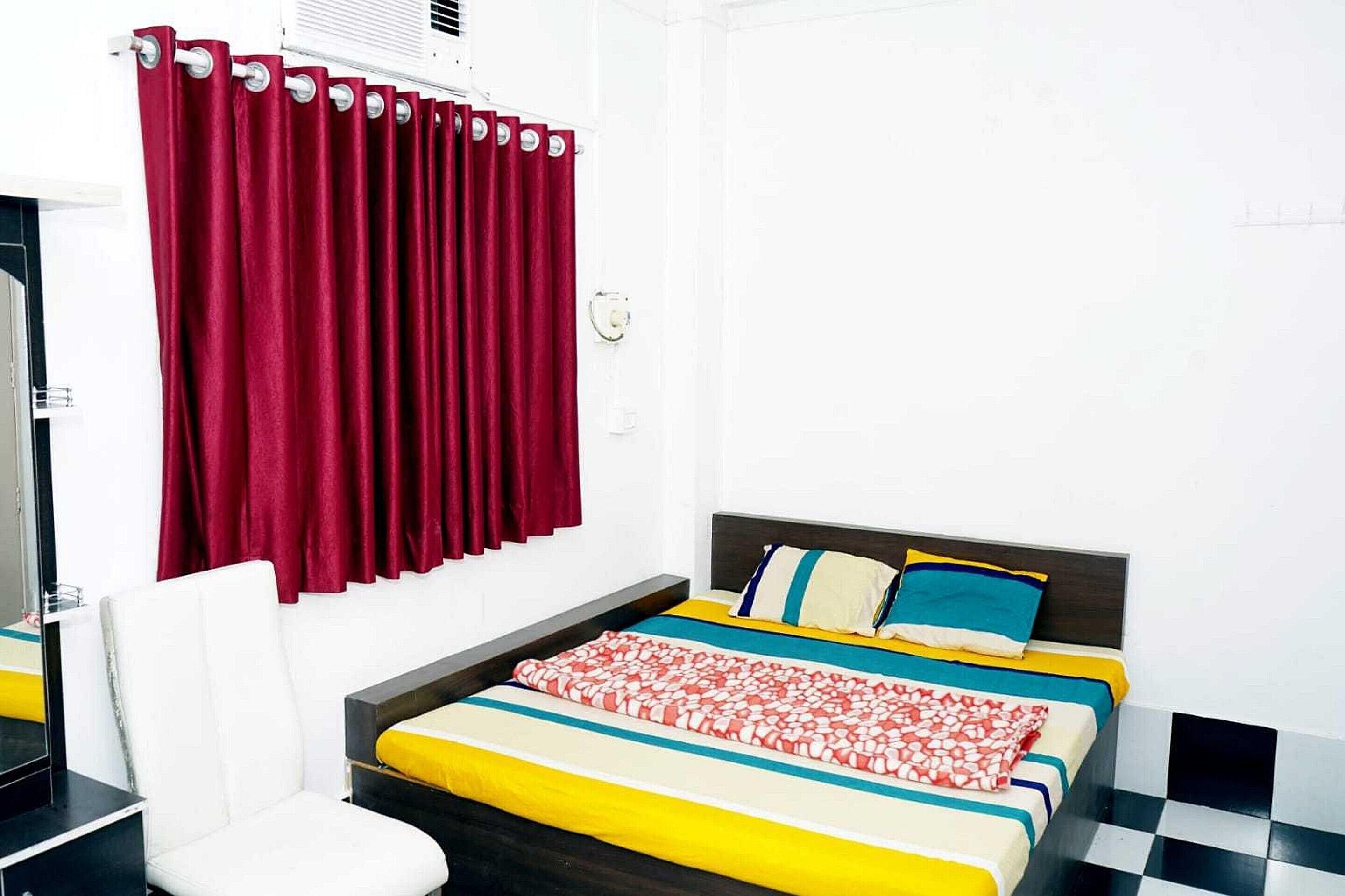 A bedroom with a bed, a chair, a window with red curtains, and a white wall.